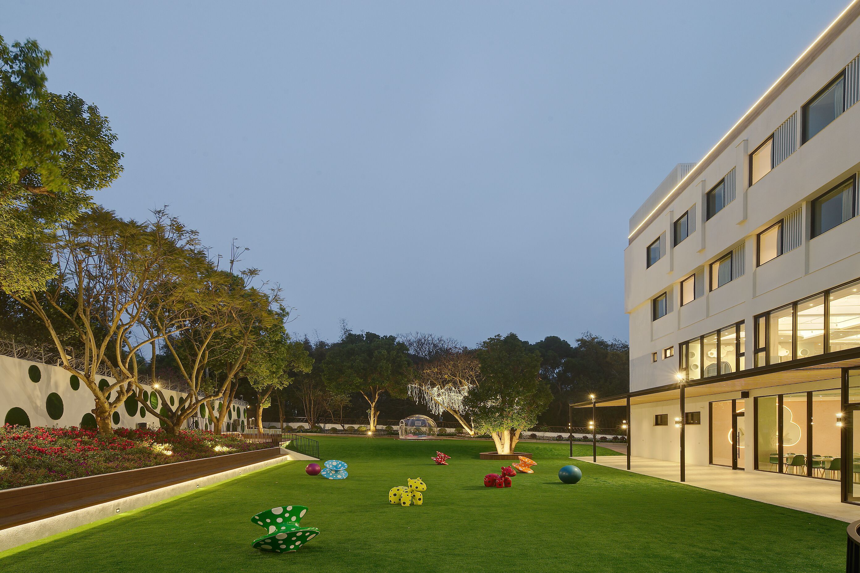 A modern building with a courtyard featuring a well-manicured lawn, colorful sculptures, and a variety of trees.