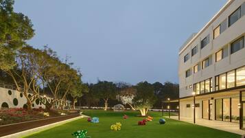 A modern building with a courtyard featuring a well-manicured lawn, colorful sculptures, and a variety of trees.