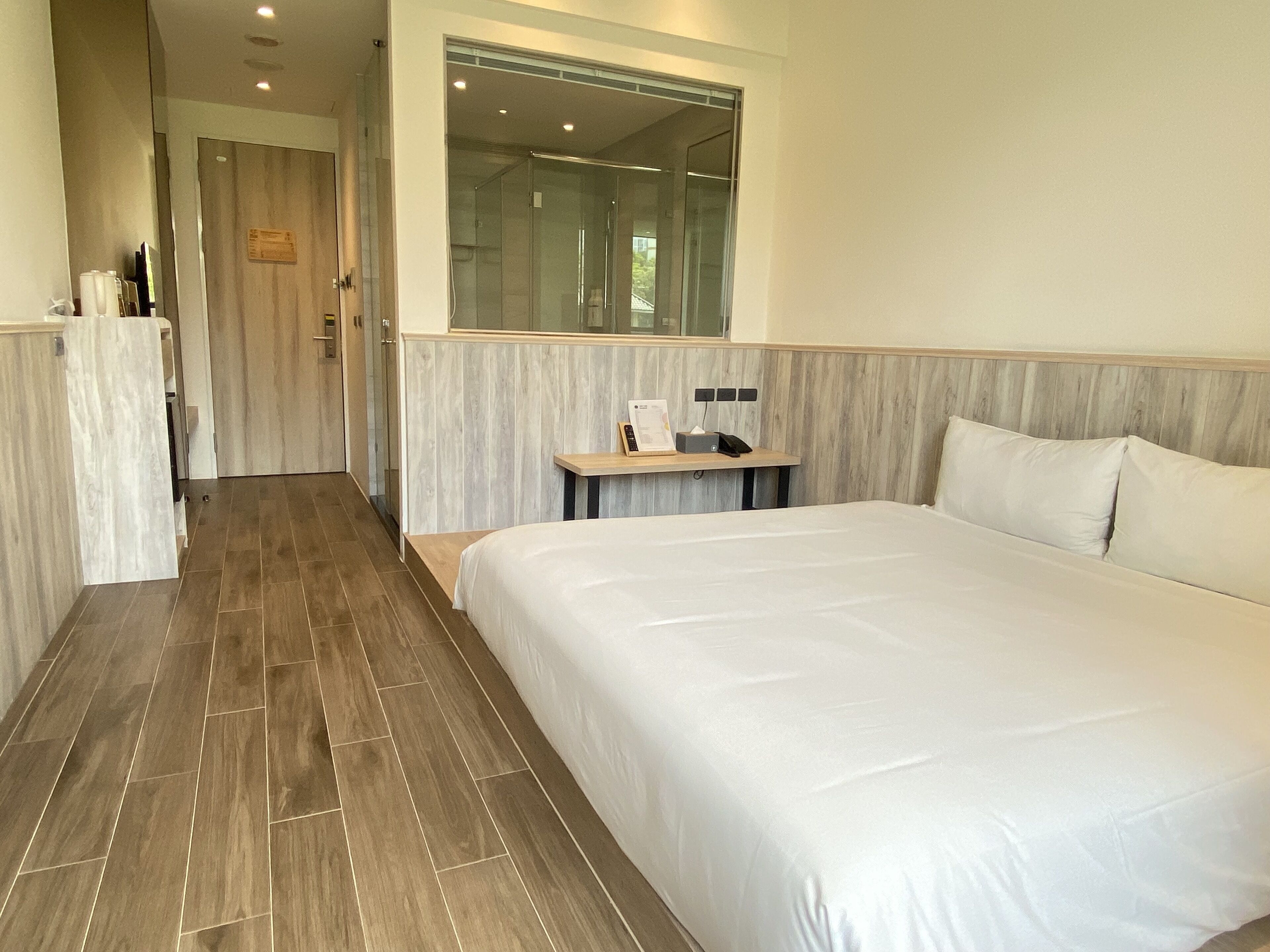 A hotel room with a large bed, a bedside table with a lamp, a mirror above the bed, and a shower area visible through a glass door.