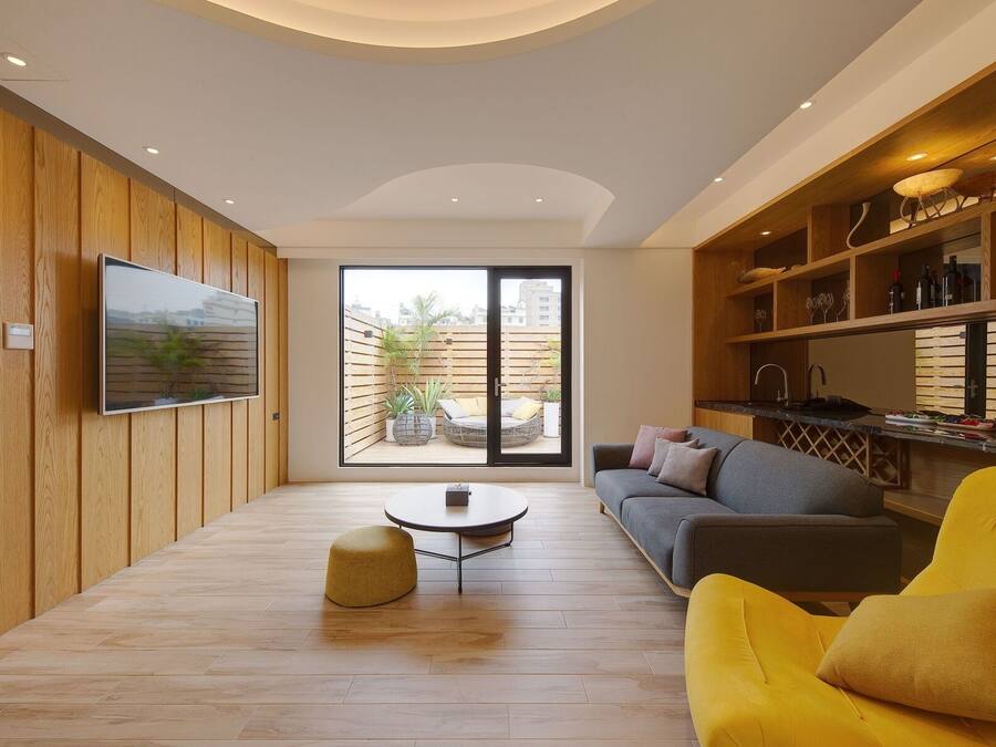 Una sala de estar moderna con piso de madera, una mesa redonda para café, un sofá y un sillón amarillo. Hay un ventanal con vista a un jardín y edificios.
