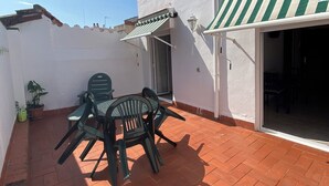Een terras met een tafel en stoelen, een potplant en een gebouw met een gestreept zonnescherm.