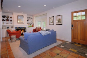 Una sala de estar con un sofá azul, un sillón rojo y una chimenea. Se ven estanterías con objetos decorativos y una puerta con un ventanal.