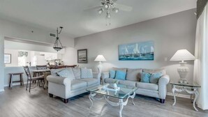 A modern living room with a glass coffee table, two sofas, a dining area with a chandelier, and a painting of sailboats on the wall.