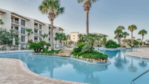 A swimming pool with a tiled deck, surrounded by palm trees and a multi-story building with balconies.