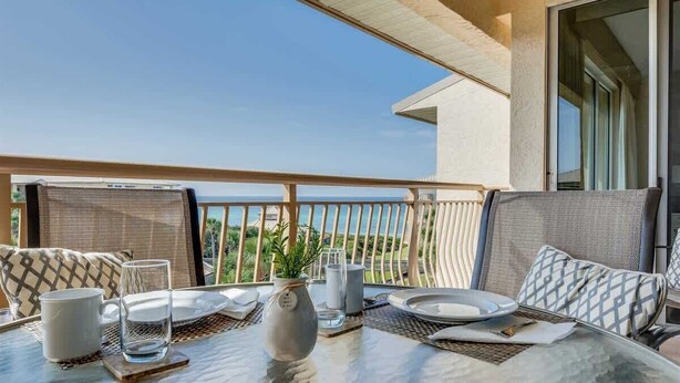 A balcony with a table set for two, featuring a glass top, two chairs, a vase, and a glass.