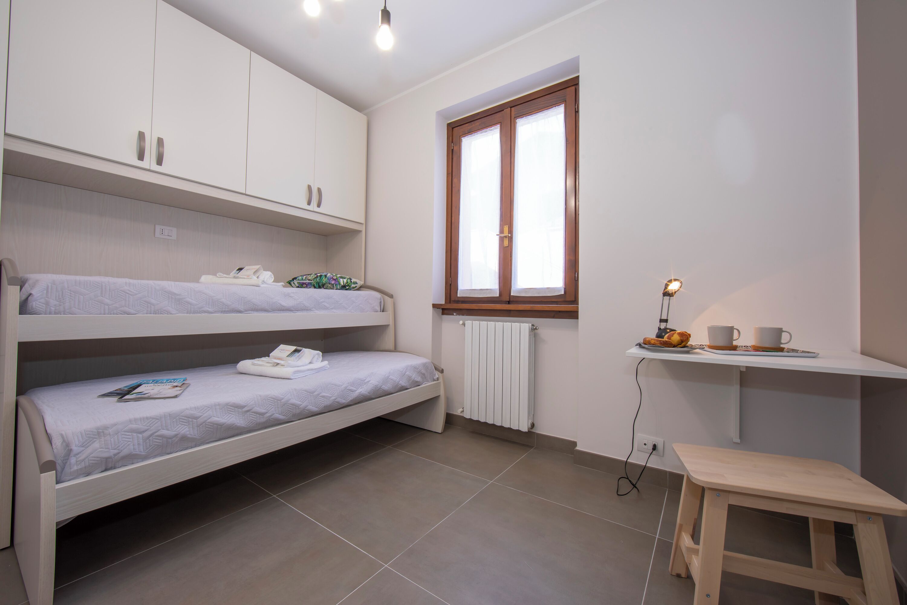 A room with bunk beds, a desk with a chair, a radiator, and a window.