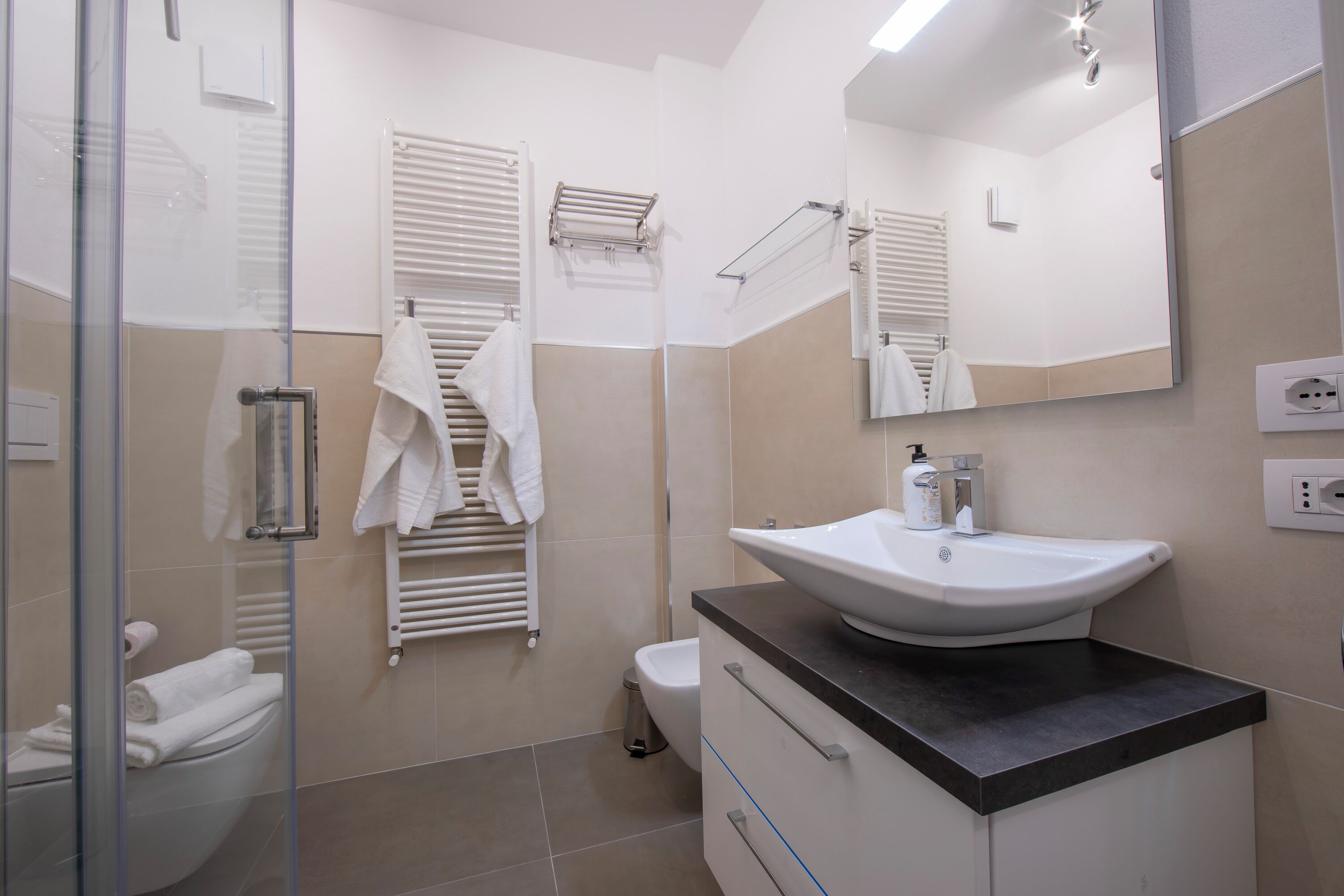 Une salle de bain moderne avec une douche à parois de verre, des toilettes blanches, un sol carrelé de couleur beige, un lavabo noir sur un plan de travail et un miroir au-dessus du lavabo.