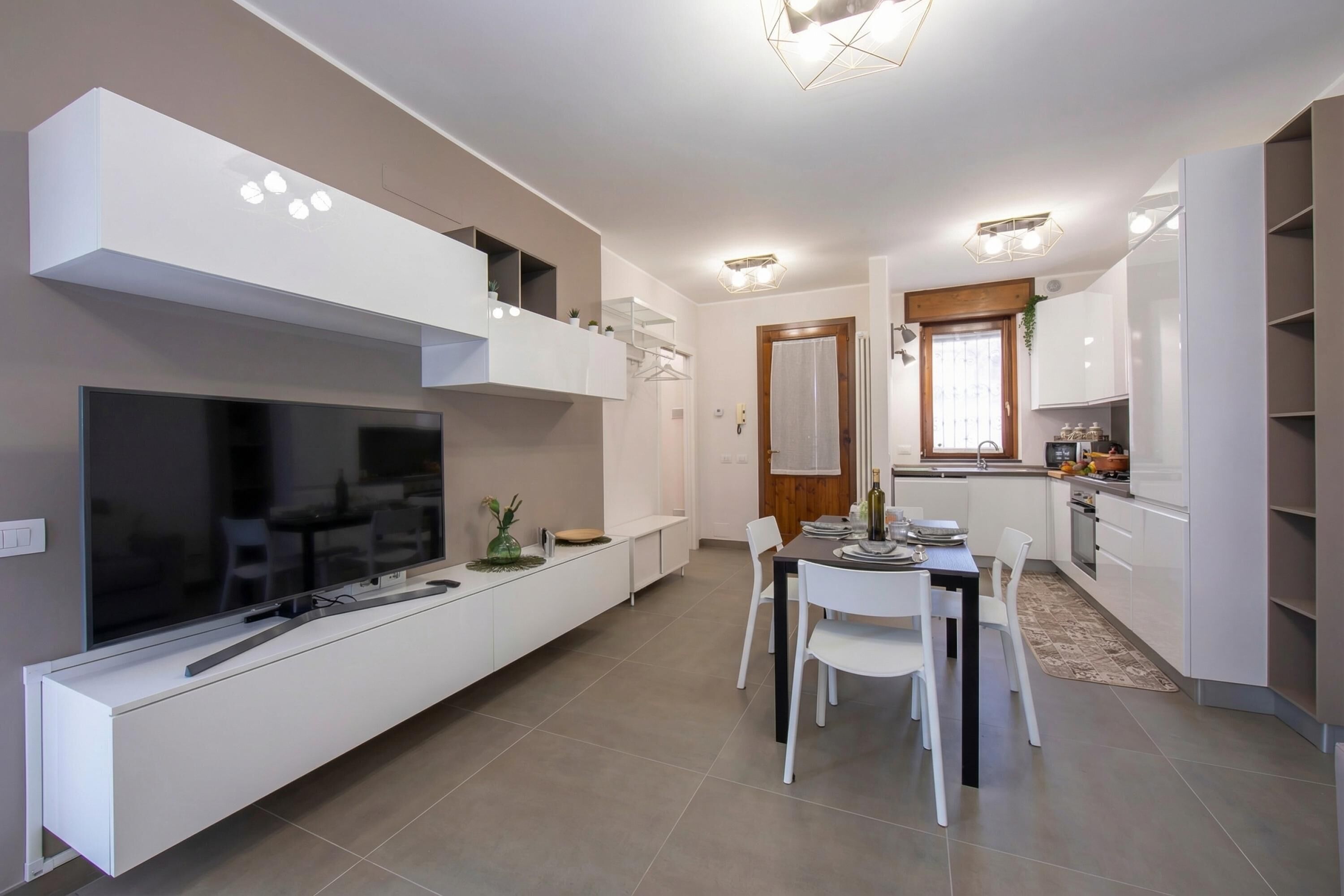 A modern kitchen with white cabinetry, a dining area with a table and chairs, a built-in TV, and a ceiling light fixture.