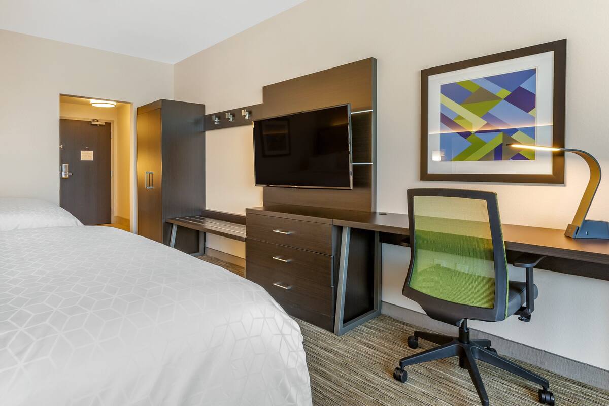 A hotel room with a bed, a desk with a chair, a TV, and a framed picture on the wall.