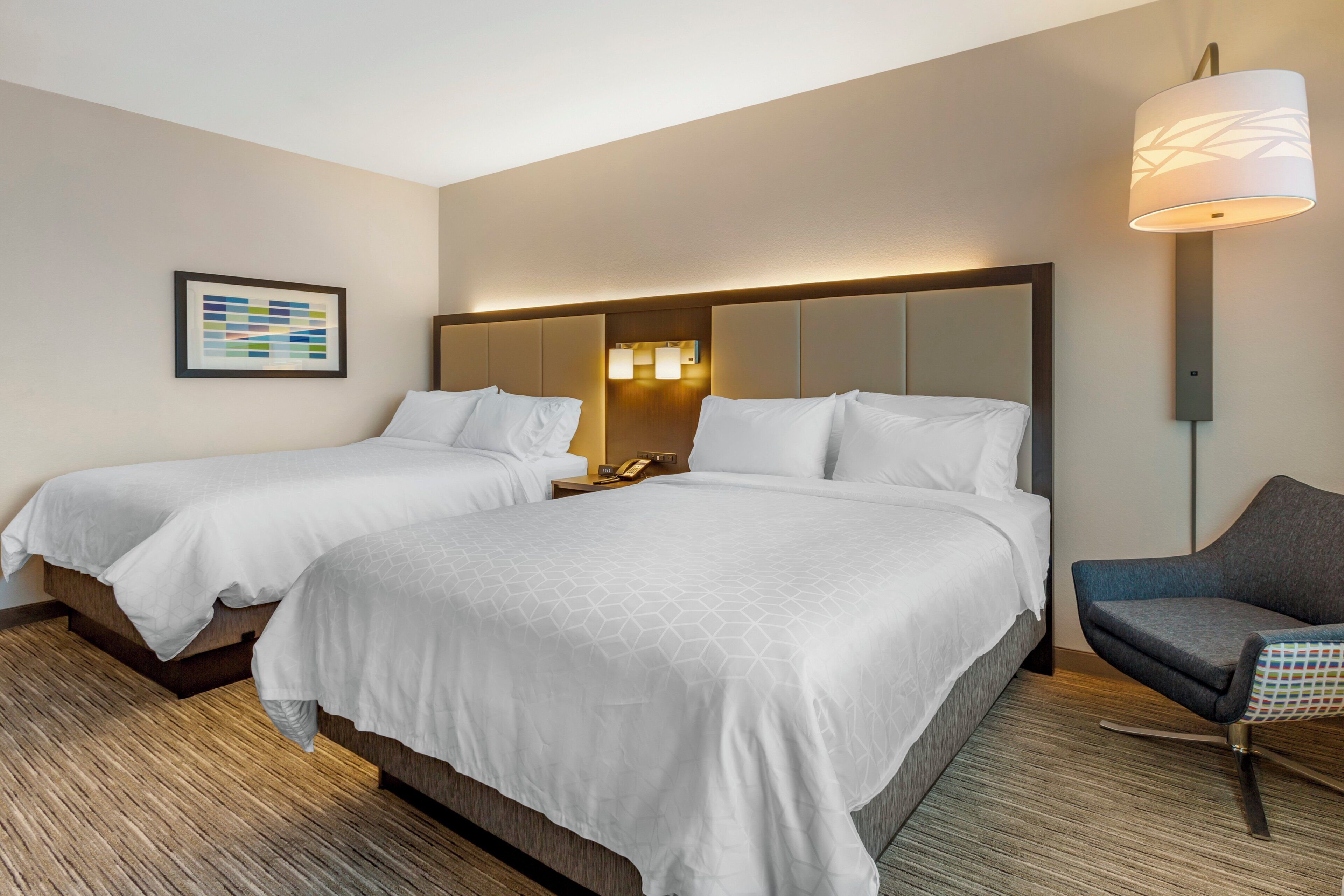 A hotel room with two beds, a bedside table with a phone, a lamp, and a chair.