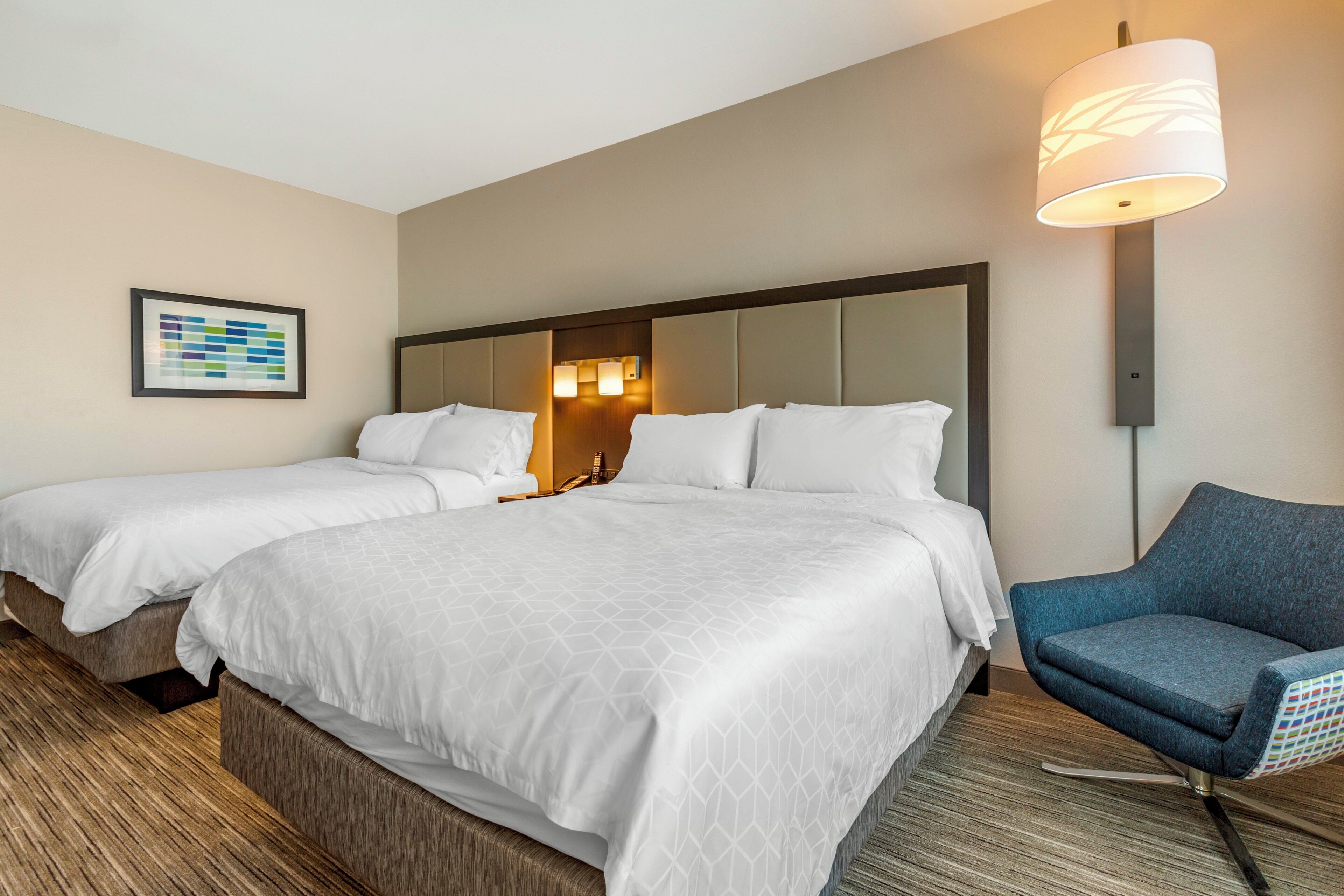 A hotel room with two beds, a blue armchair, a framed picture on the wall, and a lamp.