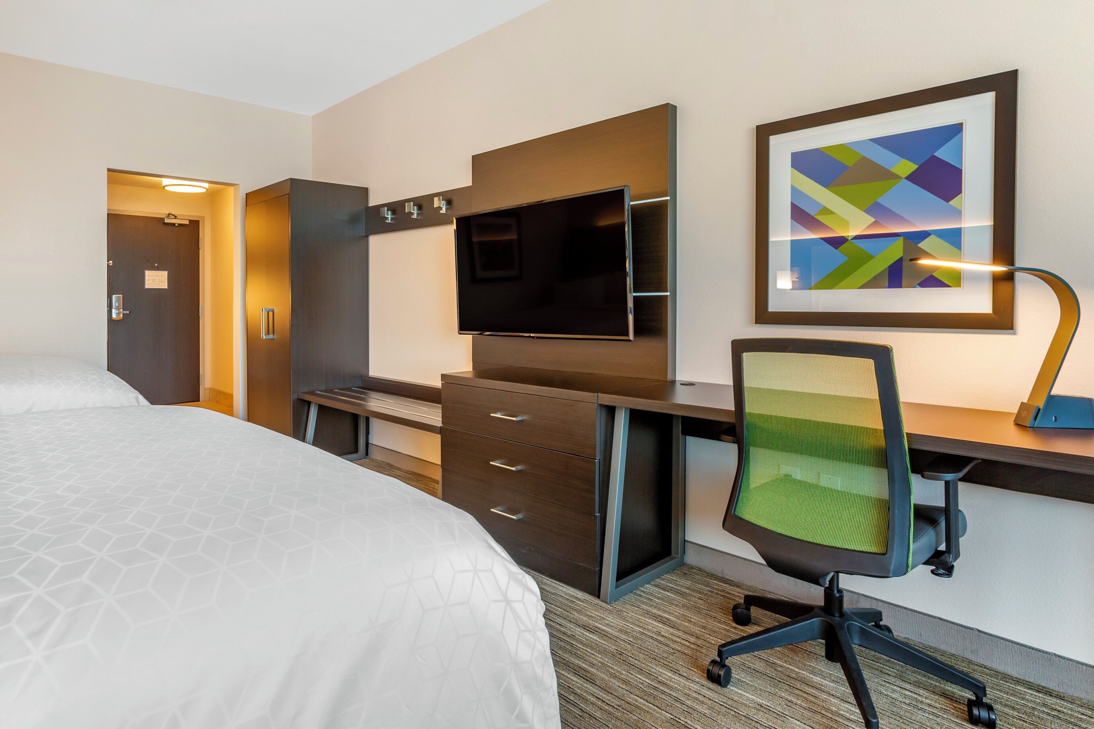 A hotel room with a bed, a desk with a chair, a television, and a framed artwork on the wall.