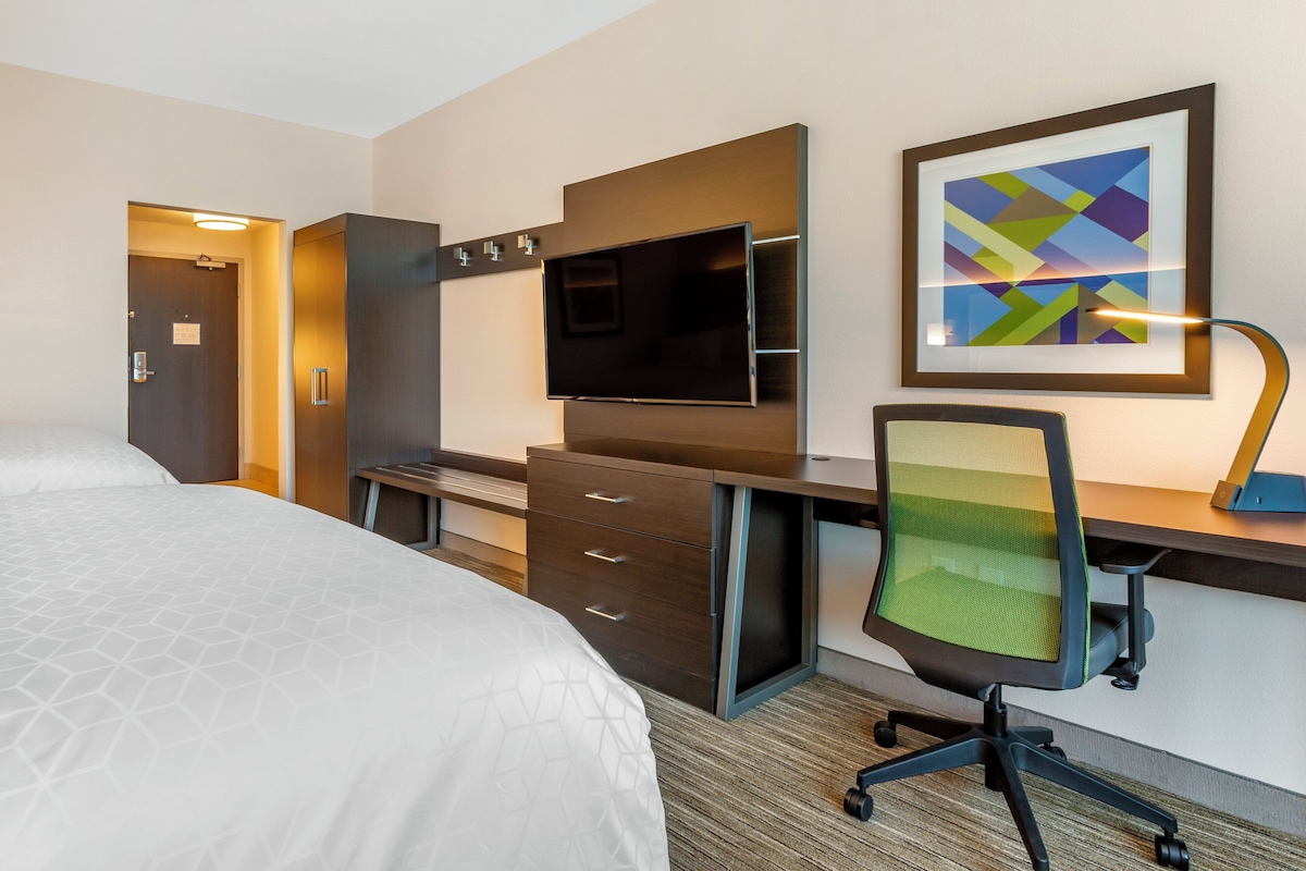 Ein Hotelzimmer mit einem Bett, einem Schreibtisch mit Stuhl, einem Fernseher und einem gerahmten Kunstwerk an der Wand.