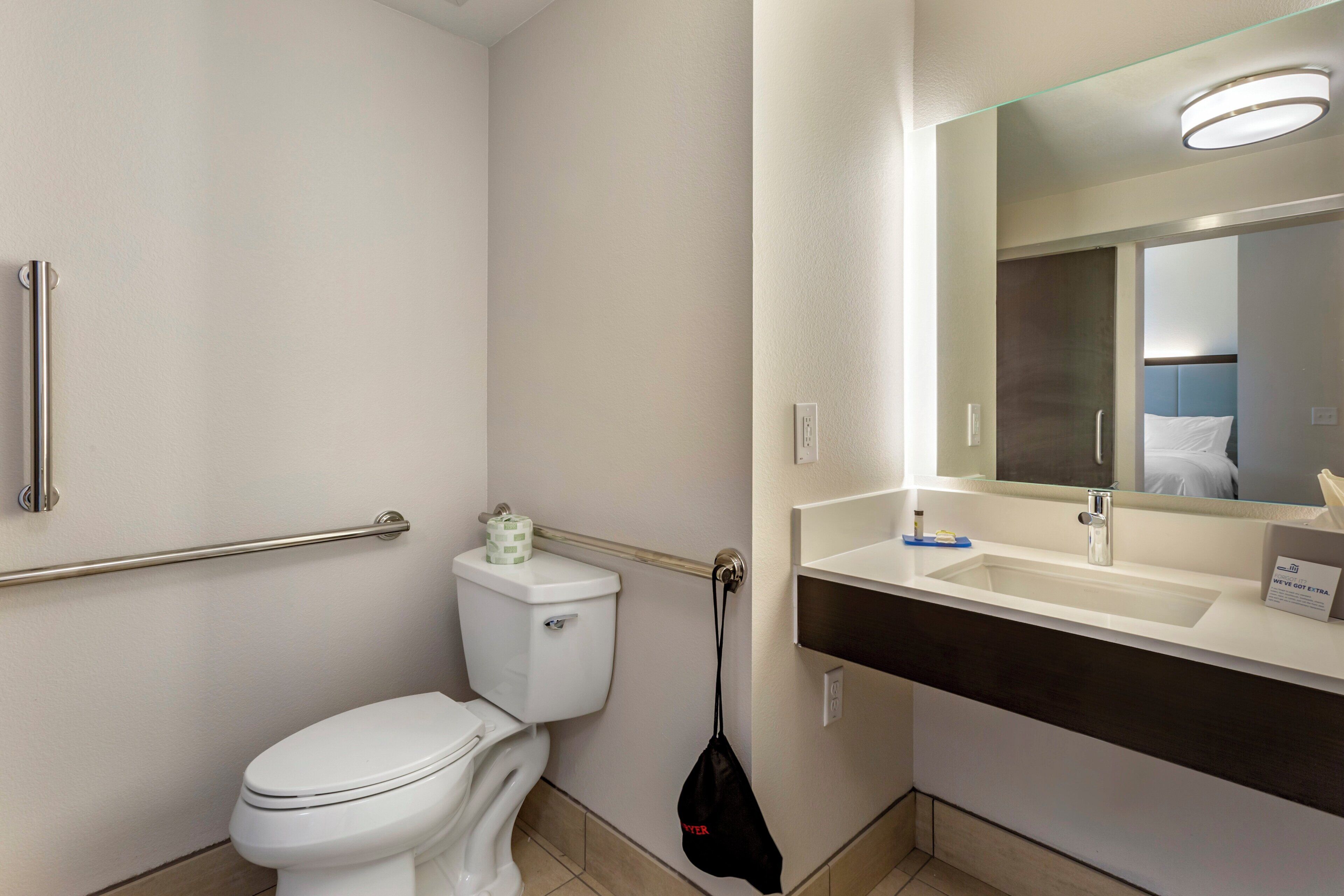Une salle de bain avec des toilettes blanches, une barre d’appui, un lavabo et un miroir.