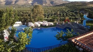 Une piscine avec un toboggan, entourée de verdure et avec une chaîne de montagnes en arrière-plan.