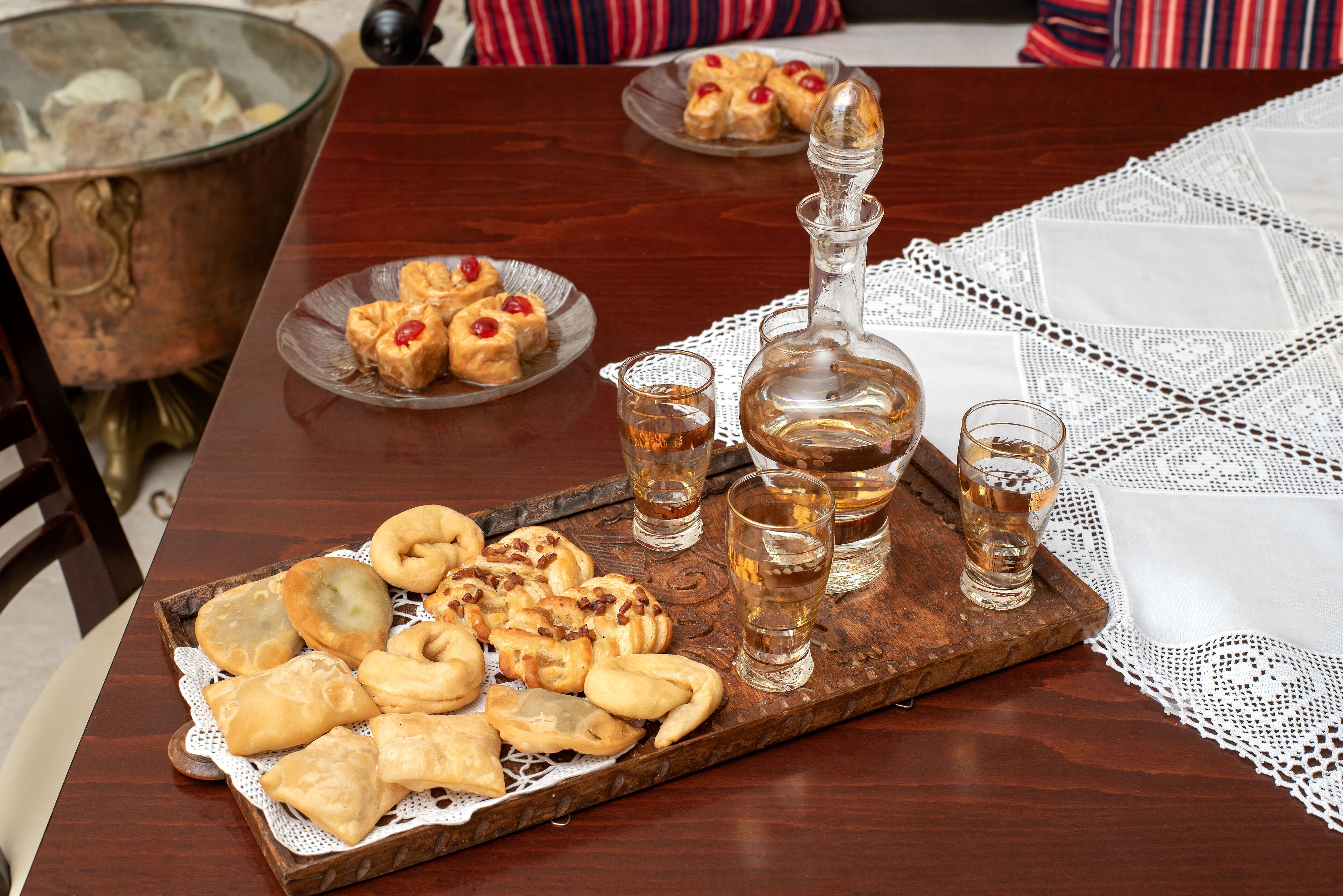 A wooden table with a tray of pastries and glasses of a clear liquid, possibly tea or whiskey, on a lace tablecloth.