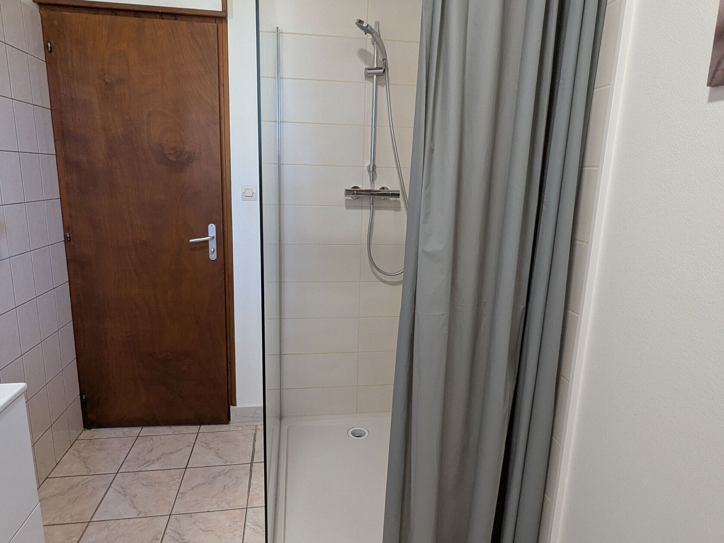 A bathroom with a shower, a wooden door, and tiled flooring.