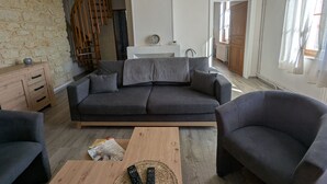 Un salon chaleureux avec un canapé gris foncé, une table basse en bois et un fauteuil gris. Un escalier en bois mène à une mezzanine, et une horloge est fixée au mur.