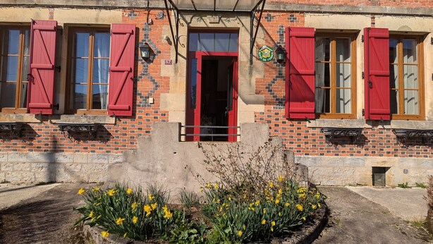 Un bâtiment en briques avec des volets rouges, une enseigne verte portant l’inscription « Ville de France » et le numéro 57, et un parterre de fleurs jaunes.