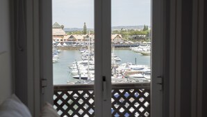 A view from a window overlooking a marina with boats docked, a building with a distinctive roof, and a distant landscape. - Marina Pearl - Vilamoura (Vilamoura)