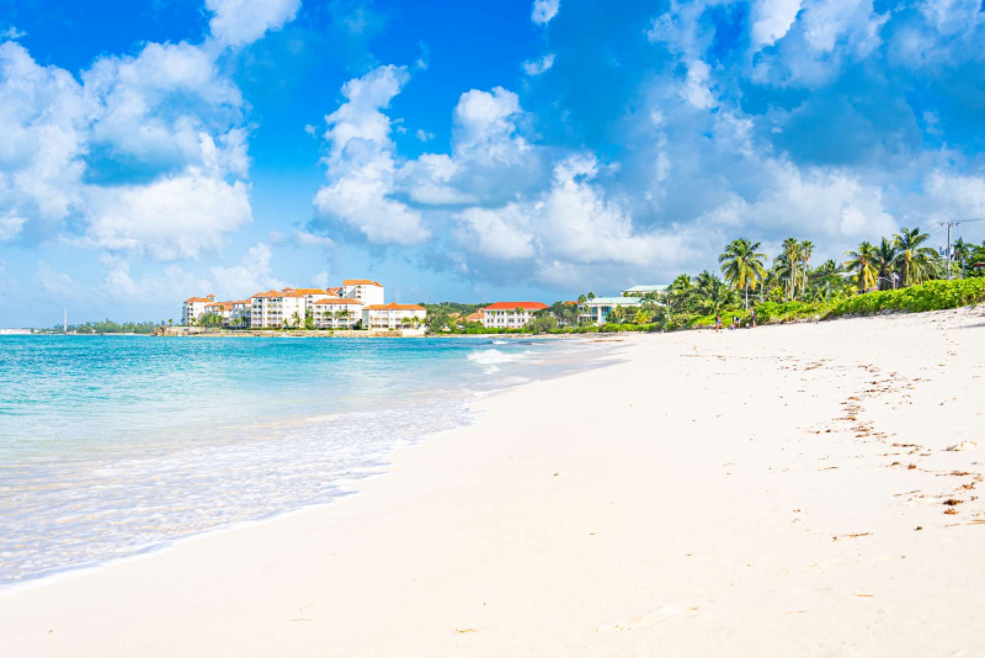 A beach with a clear sky, a sandy shore, and buildings with red roofs in the background.