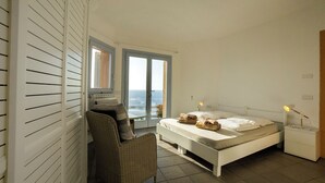 A bedroom with a bed, a wicker chair, a table, and a view of the sea through a glass door. - Exclusive clifftop villa in the heart of Gallura with infinity pool (Trinità d'Agultu e Vignola)