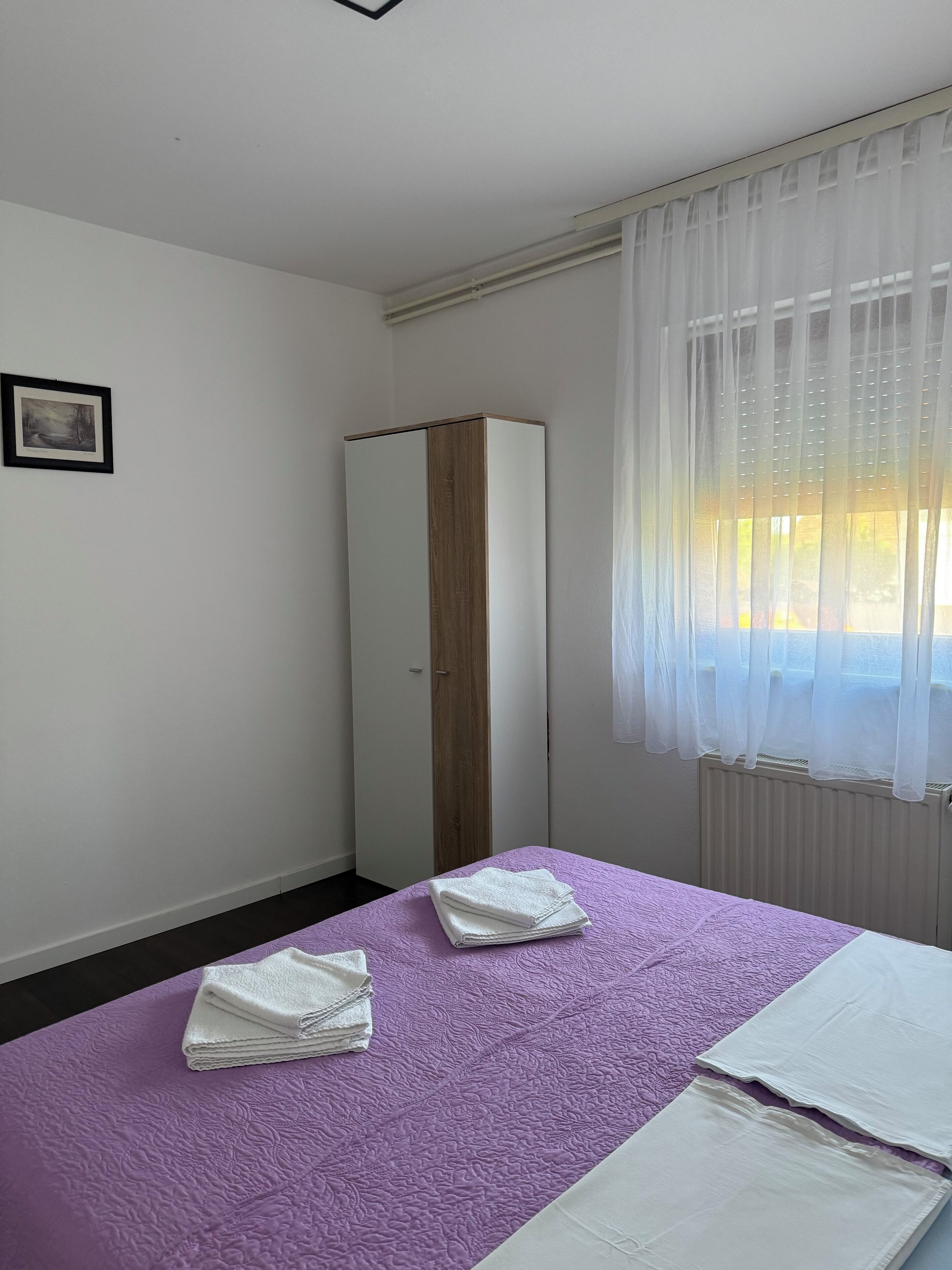 Habitación de hotel con cama, armario, ventana con cortinas traslúcidas y un cuadro enmarcado en la pared.