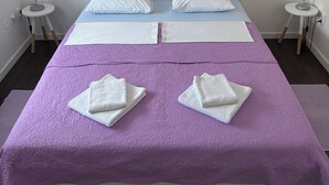 A neatly made bed with a purple bedspread, two white pillows, and two folded white towels. There are two bedside tables with lamps, and the room has white walls and dark wooden flooring.