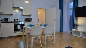 A modern kitchen with white cabinets, a microwave, and an oven. There is a dining table with chairs and a blue table runner. A TV is mounted on the wall, and a window with blue curtains is visible. - Jimmy's Apartments Suites (Vienna)