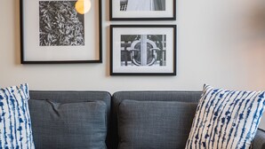 A grey sofa with two blue and white patterned pillows, a white round side table with a remote control, and three framed pictures on the wall. - Jimmy's Apartments Suites (Vienna)