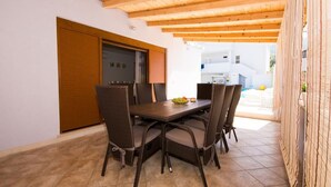 Un coin repas avec une table en bois et des chaises en osier, une porte coulissante et, en arrière-plan, une vue sur un bâtiment blanc.