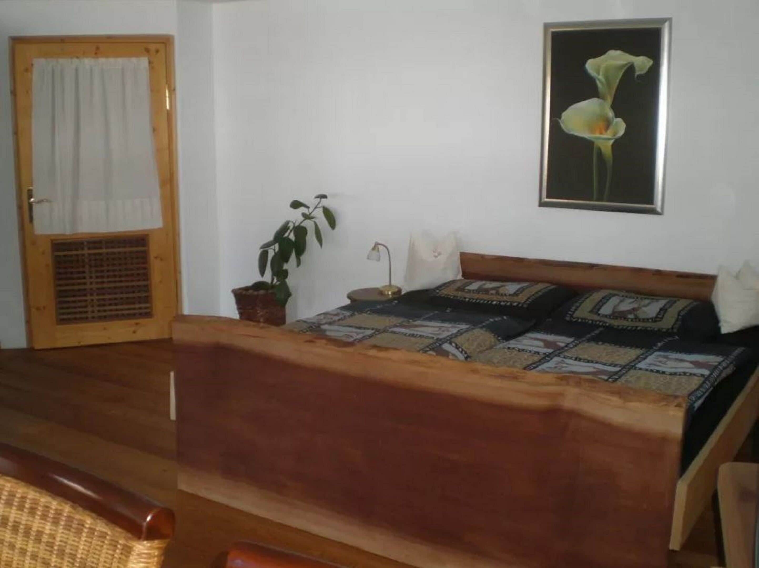 A bedroom with a wooden bed, a potted plant, a wall-mounted lamp, and a framed picture of a flower.