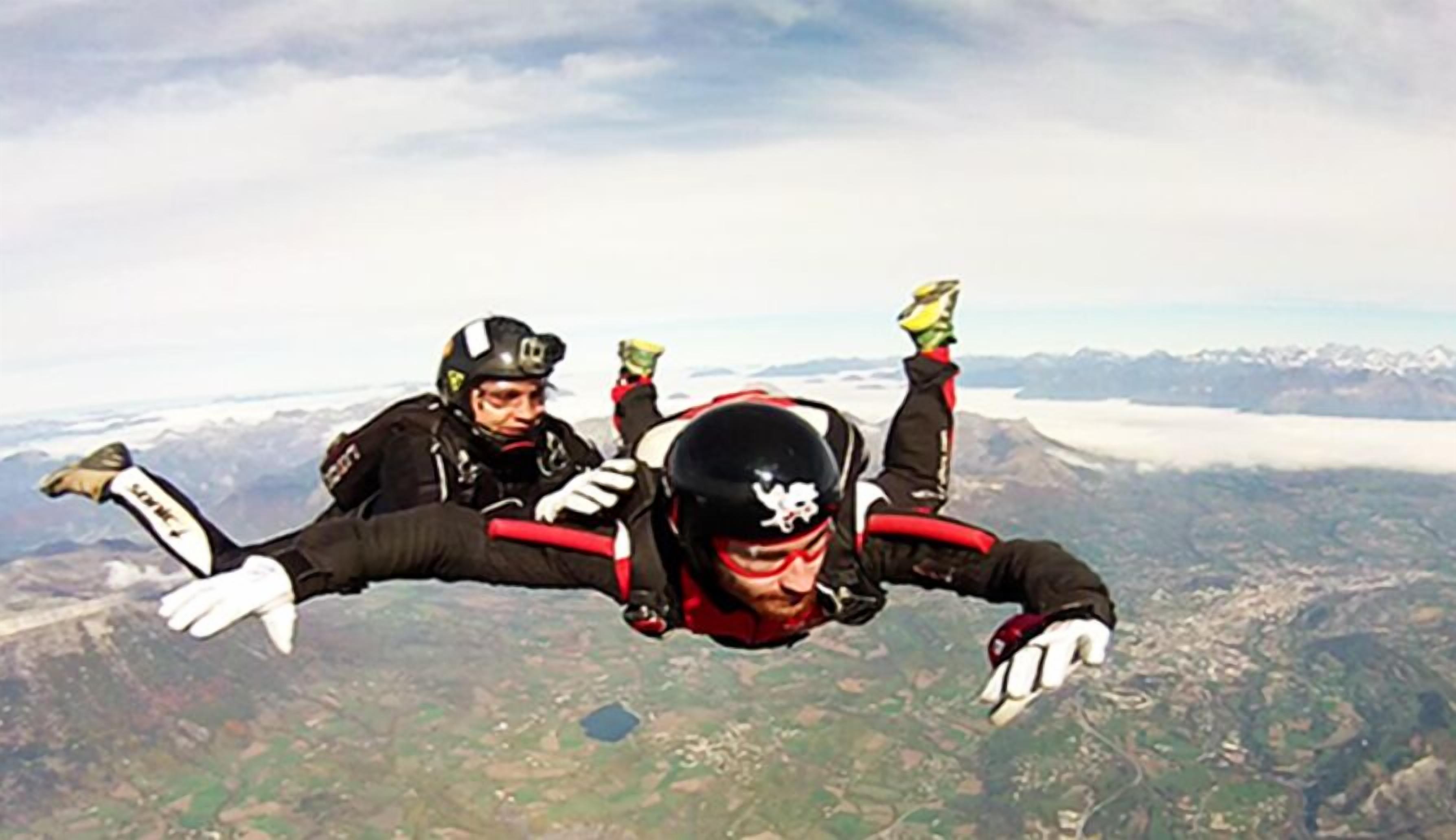 Two skydivers in freefall with a mountainous landscape below.