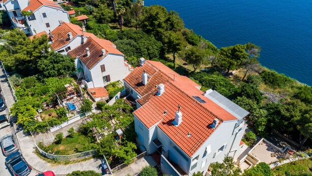Vue aérienne d’un quartier résidentiel situé sur la côte, avec des maisons aux toits recouverts de tuiles rouges et aux murs blancs, entourées de verdure et bordées par une mer bleue et limpide.