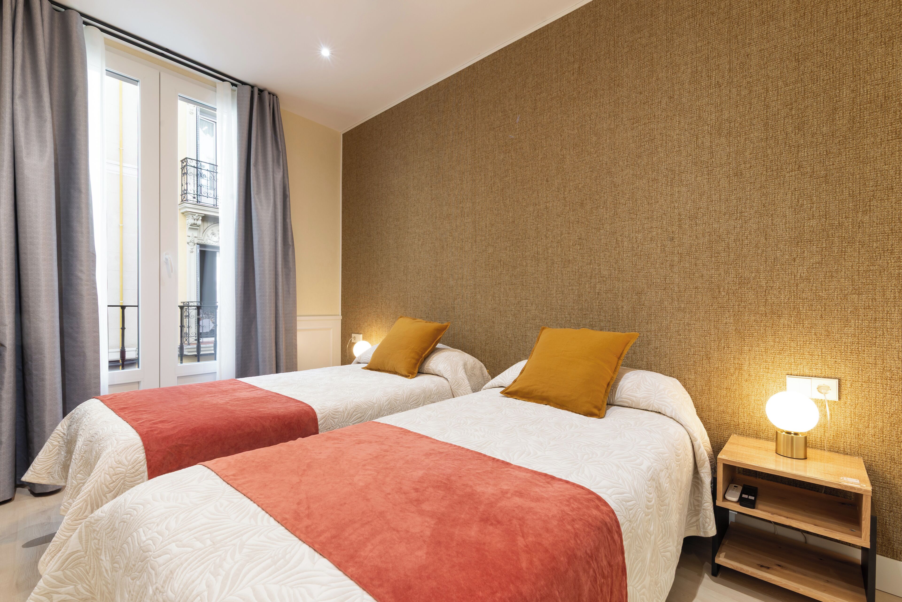 Habitación de hotel con dos camas, una mesita de noche con lámpara y vistas a un balcón.