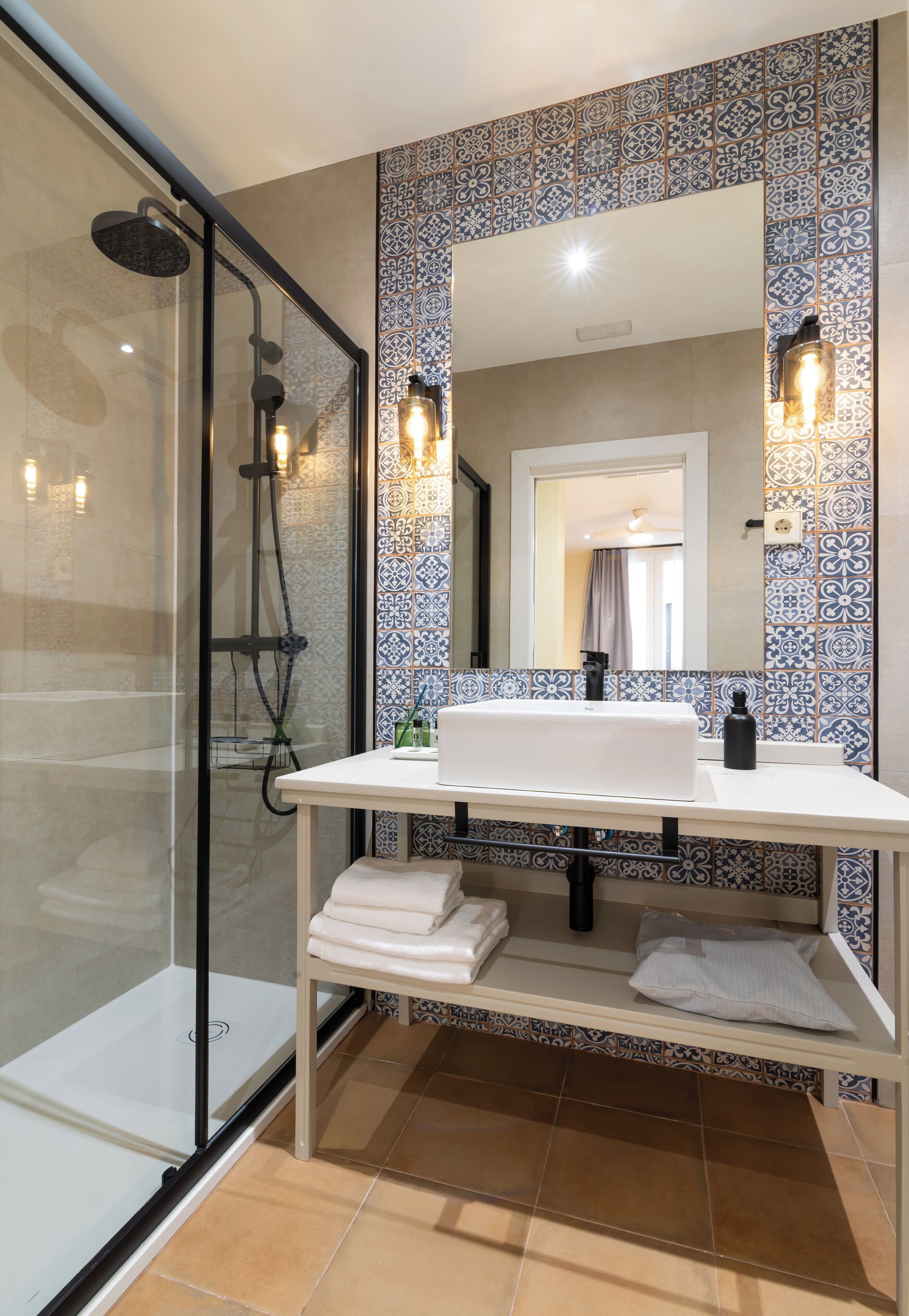 Un baño moderno con ducha envuelta en vidrio, un lavamanos blanco con espejo superior y azulejos estampados en las paredes.