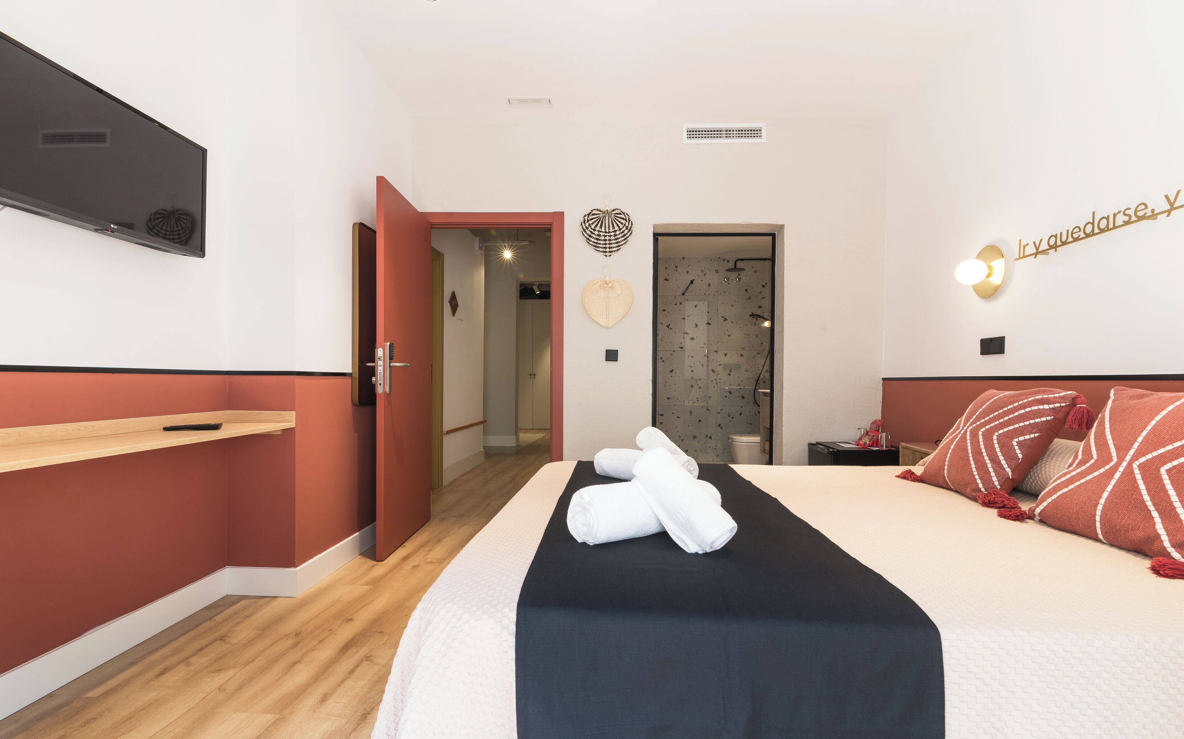 Habitación de hotel con cama, mesita de noche, televisor montado en la pared y puerta que da a un baño.