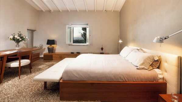A bedroom with a large bed, a desk, a chair, a nightstand, a lamp, and a window with a view of the outdoors.
