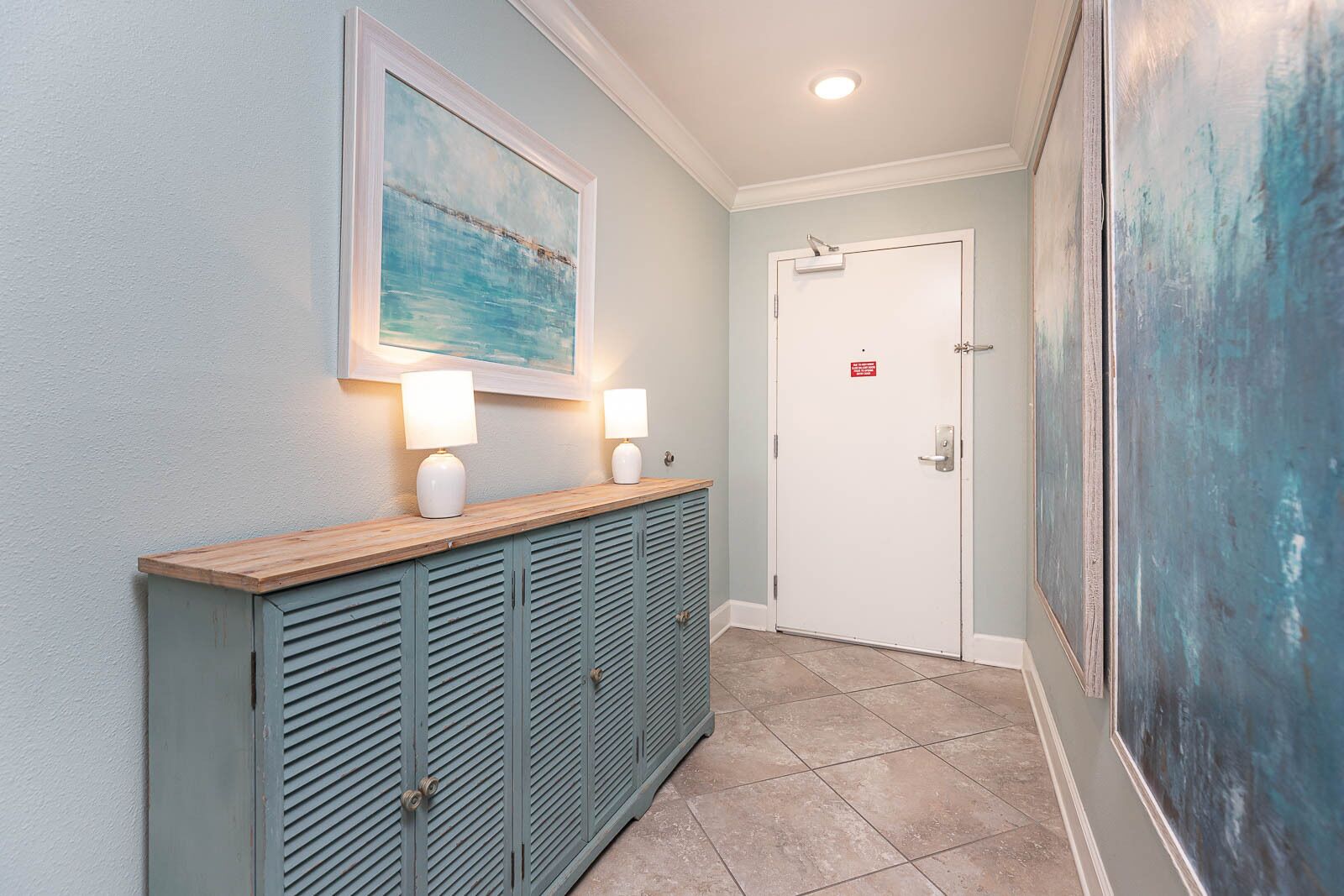 A hallway with a wooden console table, two wall-mounted lamps, a door with a sign, and a large abstract painting.