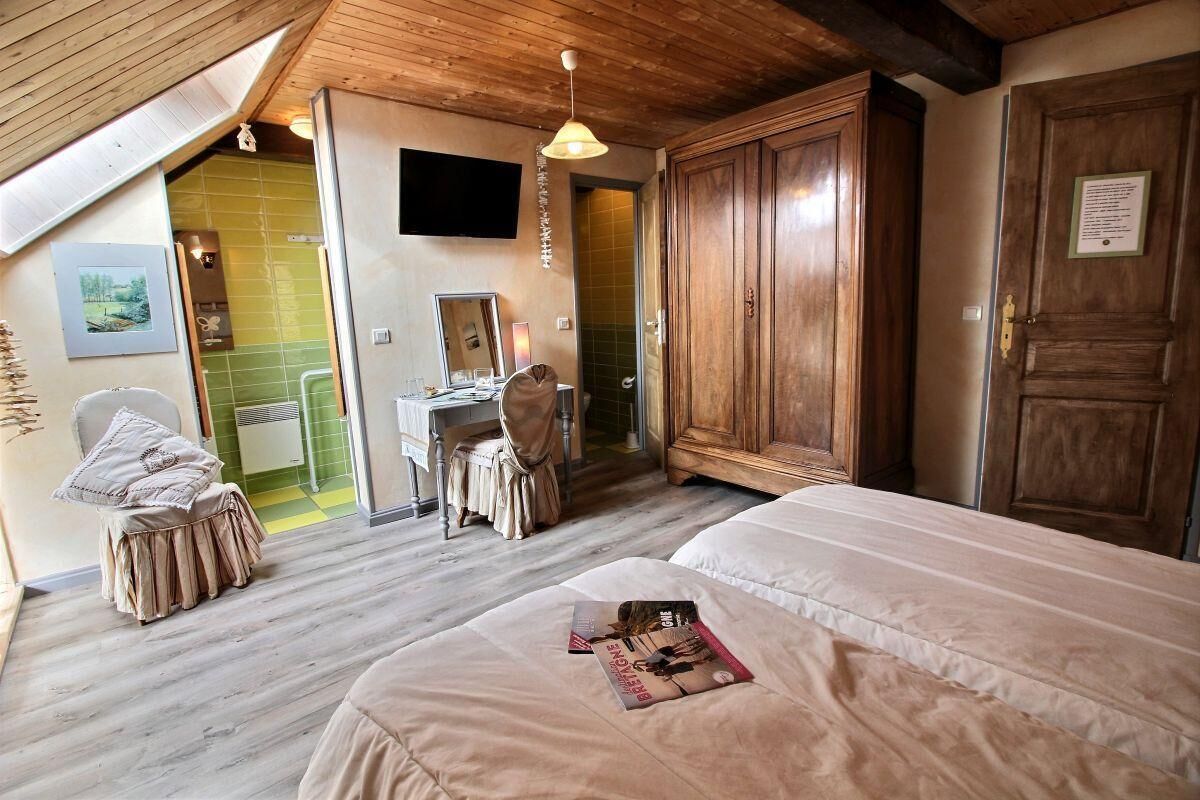 Une chambre avec deux lits, une armoire en bois, un bureau avec une chaise, et une salle de bain visible à travers une porte ouverte.