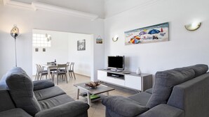 Een moderne woonkamer met grijze banken, een flatscreen televisie, een eethoek met stoelen en een schilderij van strandparasols.