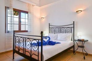 Una cama con estructura metálica, ropa de cama blanca y una toalla azul dispuesta en forma de corazón. Hay una ventana con cortinas, una mesita de noche con lámpara y un aplique de pared.