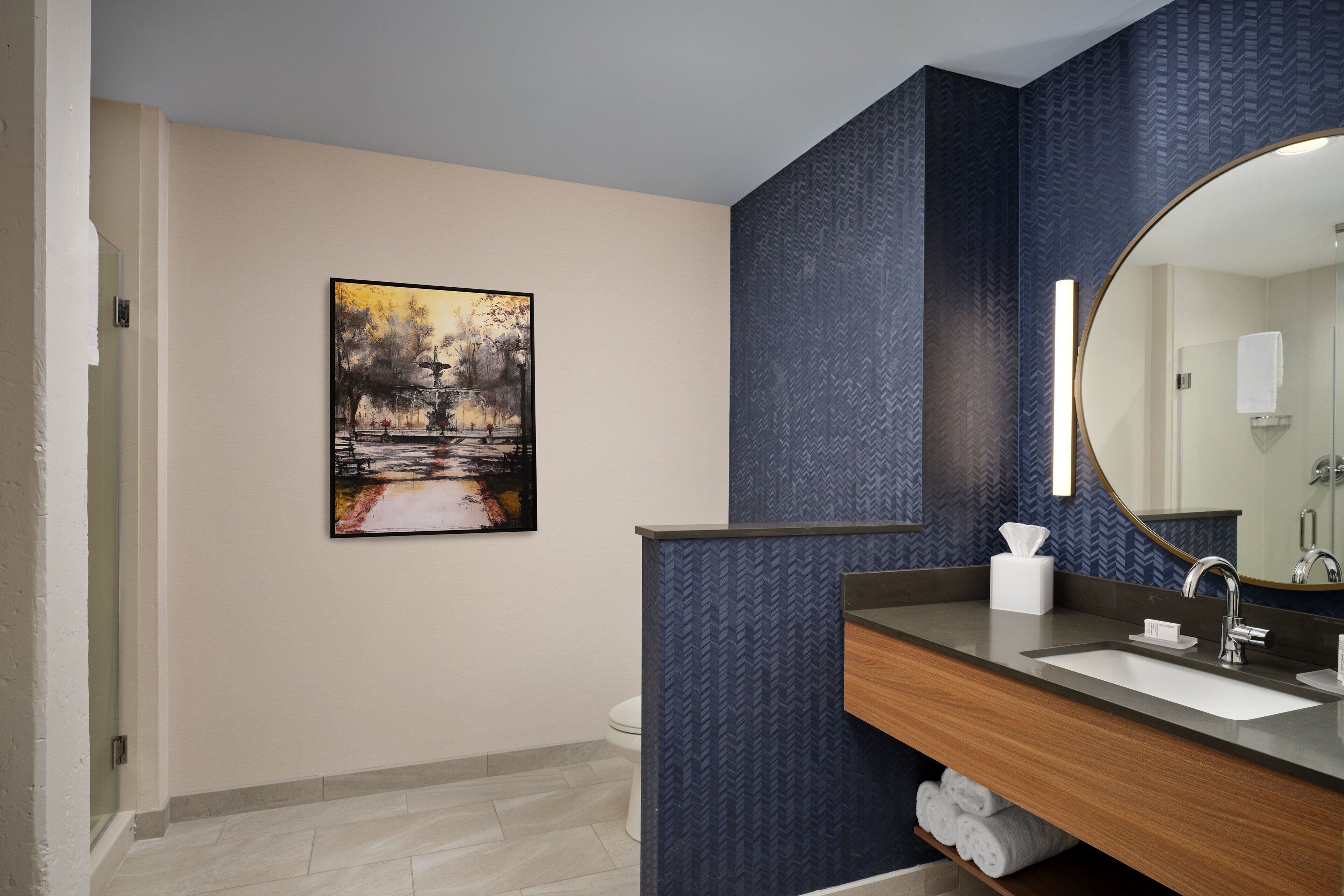 A modern bathroom with a large mirror, a wooden vanity, a sink, and a toilet. There is a painting on the wall and a towel rack under the sink.
