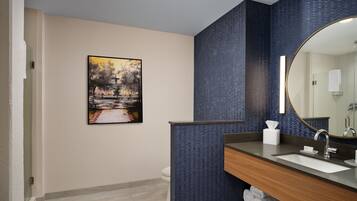 A modern bathroom with a large mirror, a wooden vanity, a sink, and a toilet. There is a painting on the wall and a towel rack under the sink.