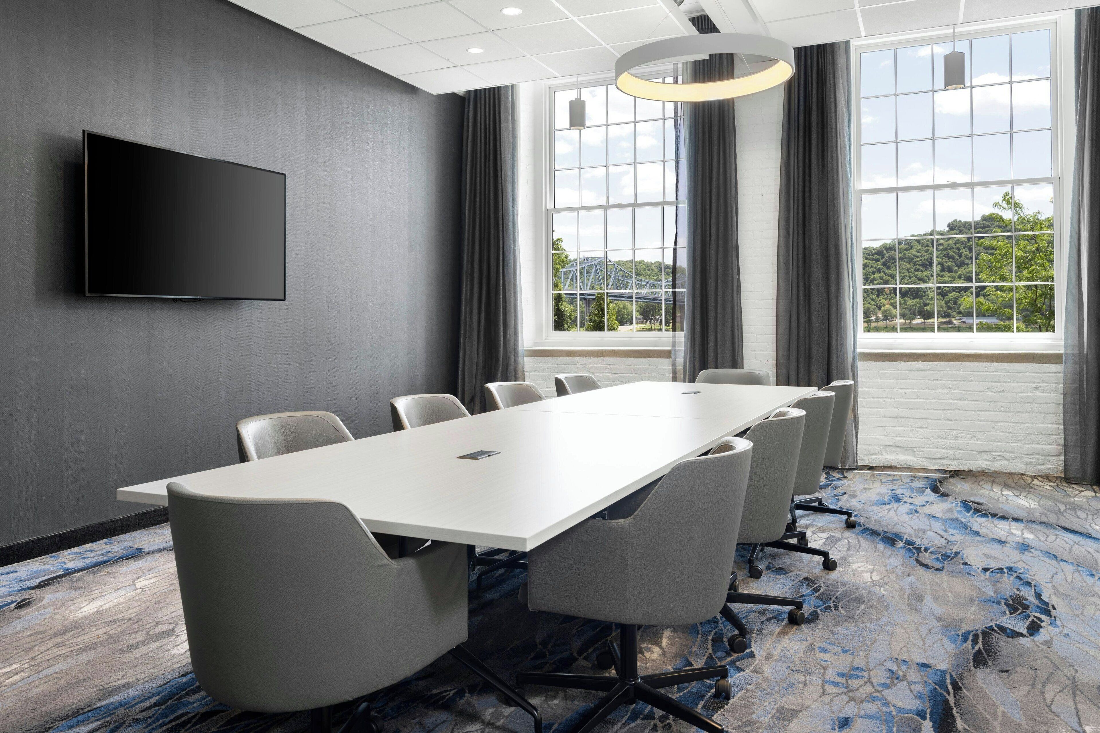 A conference room with a long table, chairs, a flat-screen TV, large windows, and a patterned carpet.