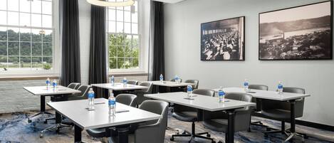 A conference room with tables, chairs, and water bottles. There are large windows with curtains and two framed pictures on the wall.