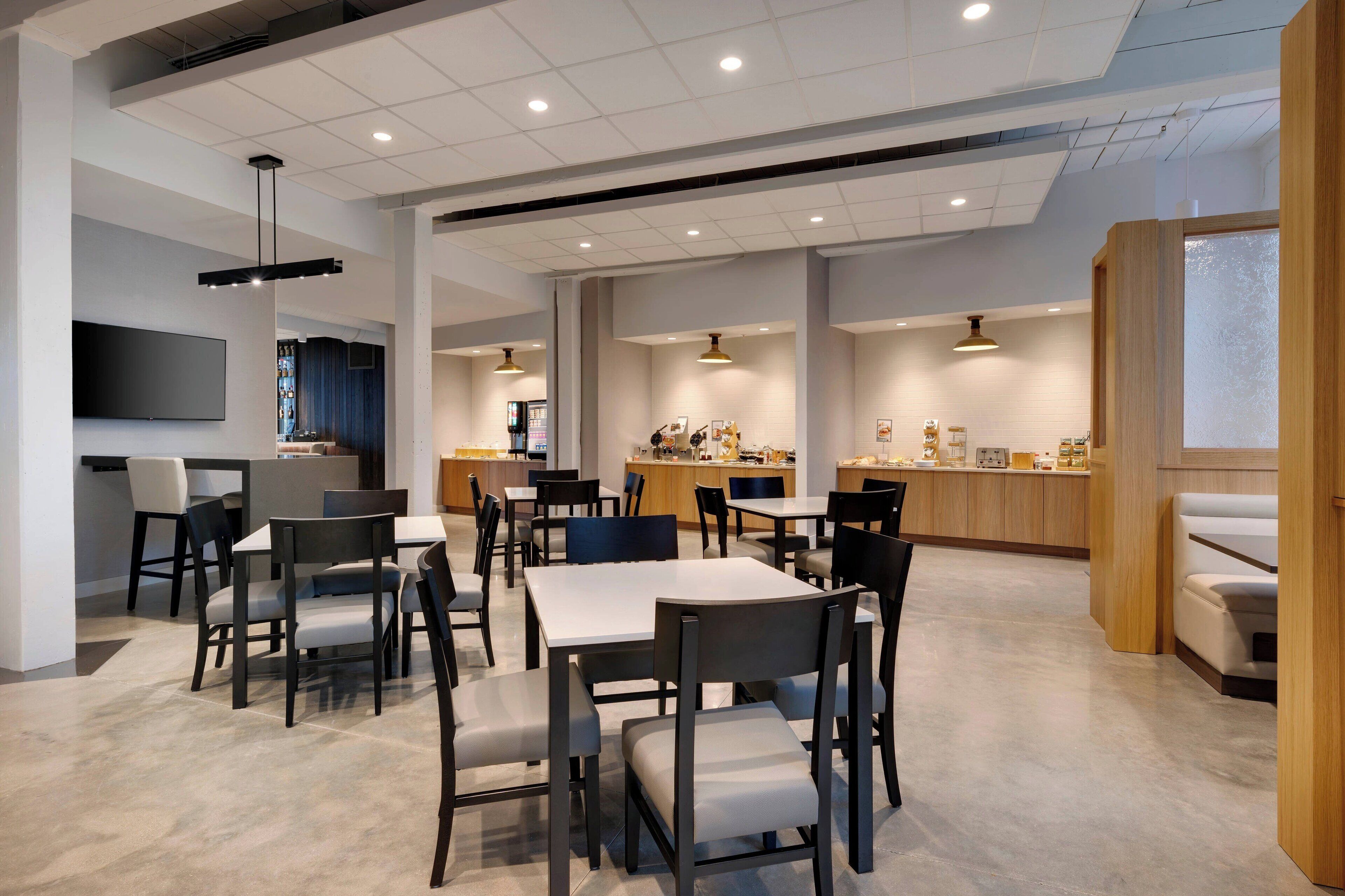 Un espace repas moderne avec des tables et des chaises, une cuisine en arrière-plan et un téléviseur à écran plat fixé au mur.