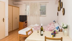 Een kleine, goed verlichte slaapkamer met een bed, een televisie, een tafel met een fles en glazen, en een raam met zonwering.