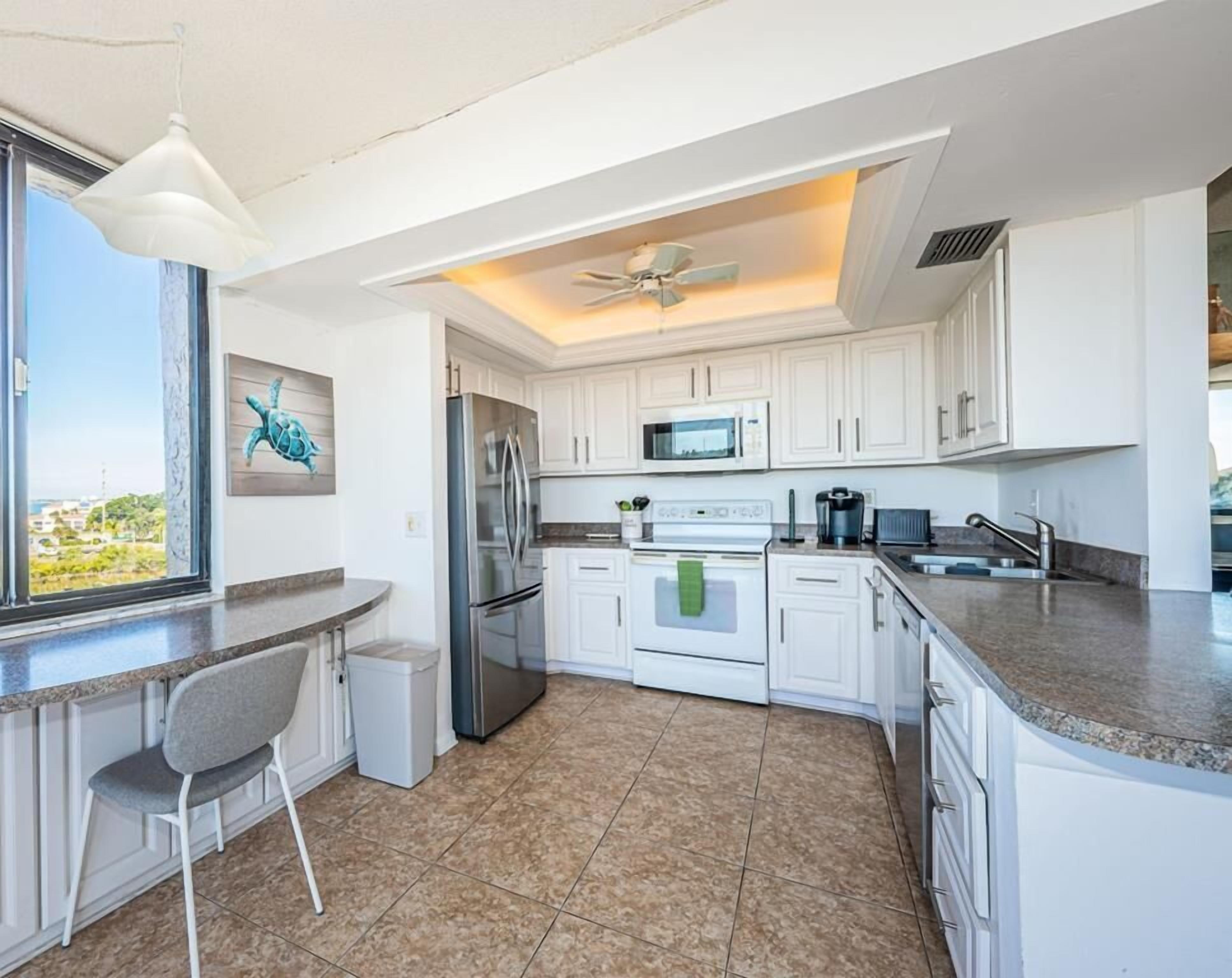 A modern kitchen with white cabinets, a stainless steel refrigerator, a microwave, and a gas stove. There is a window with a view of the outside, a ceiling fan, and a chair near the window.