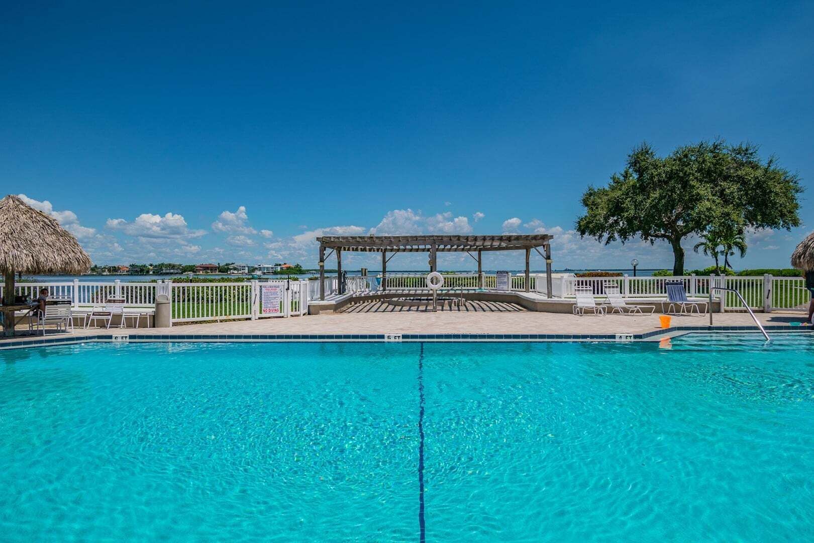 Una piscina azul cristalina con una terraza de madera, sillas de descanso y un paraguas de paja.